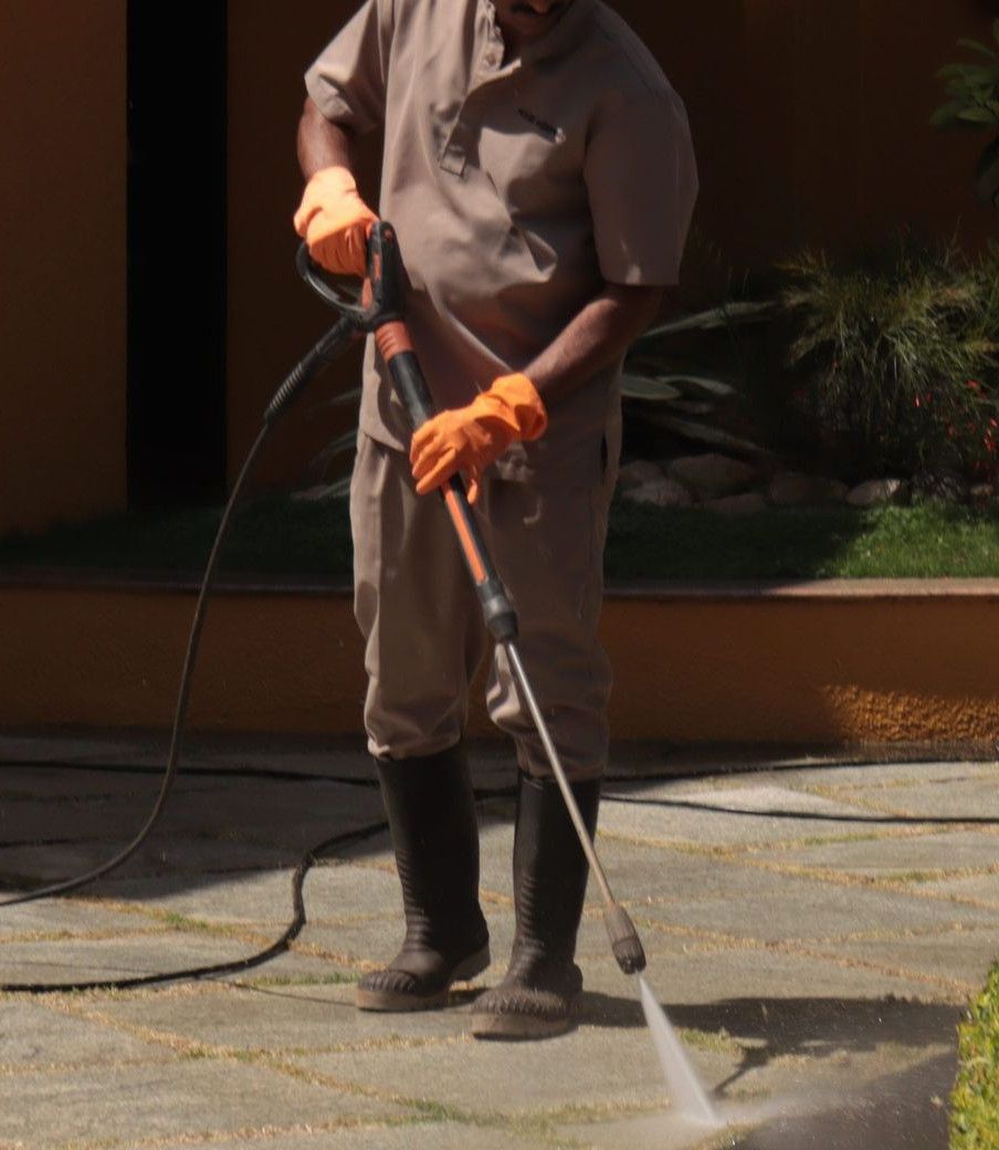 A gardener wearing a hat cleans an urban sidewalk with a pressure washer.